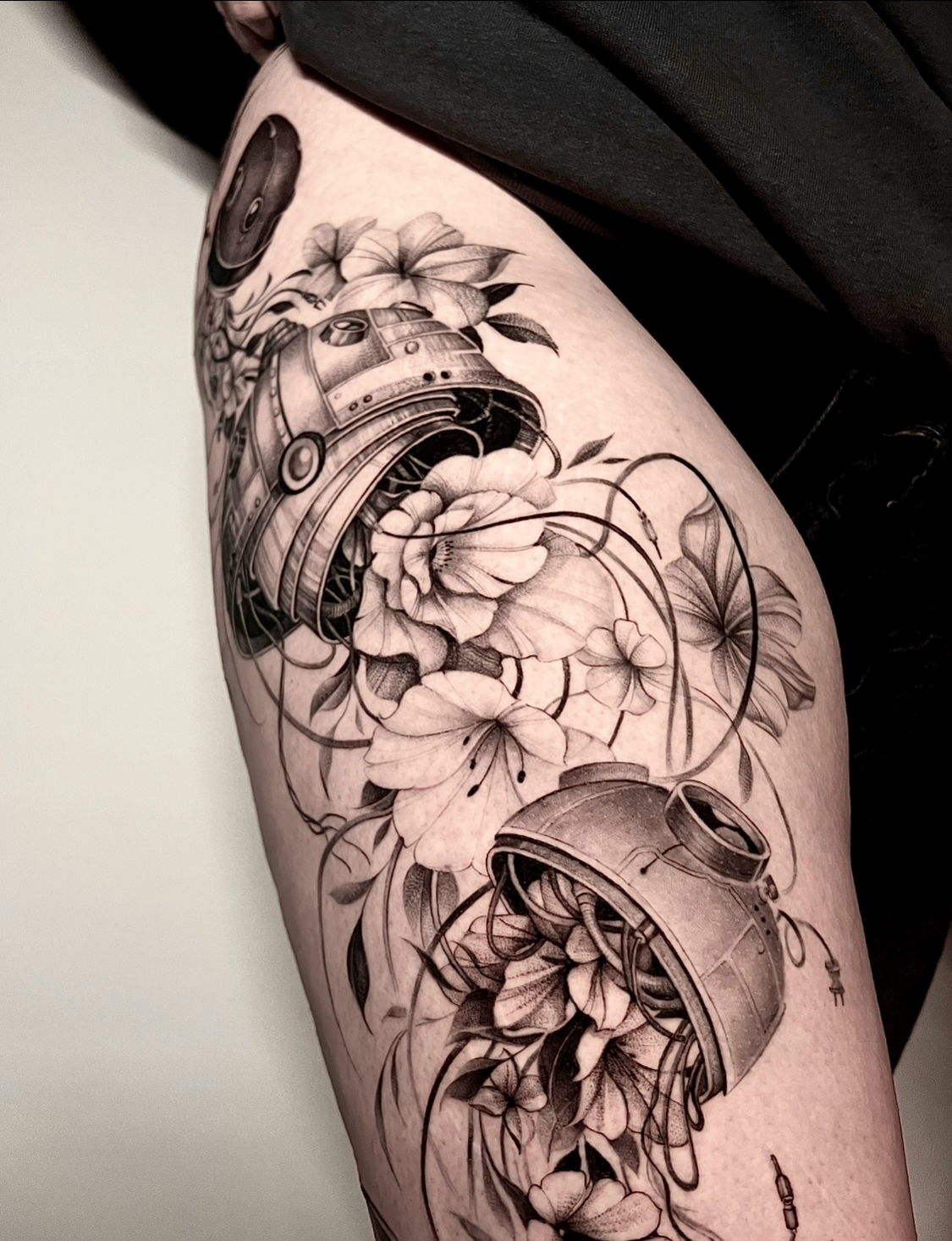 Fine line sort-grå lår-tatovering af en astromech-droide omgivet af liljer